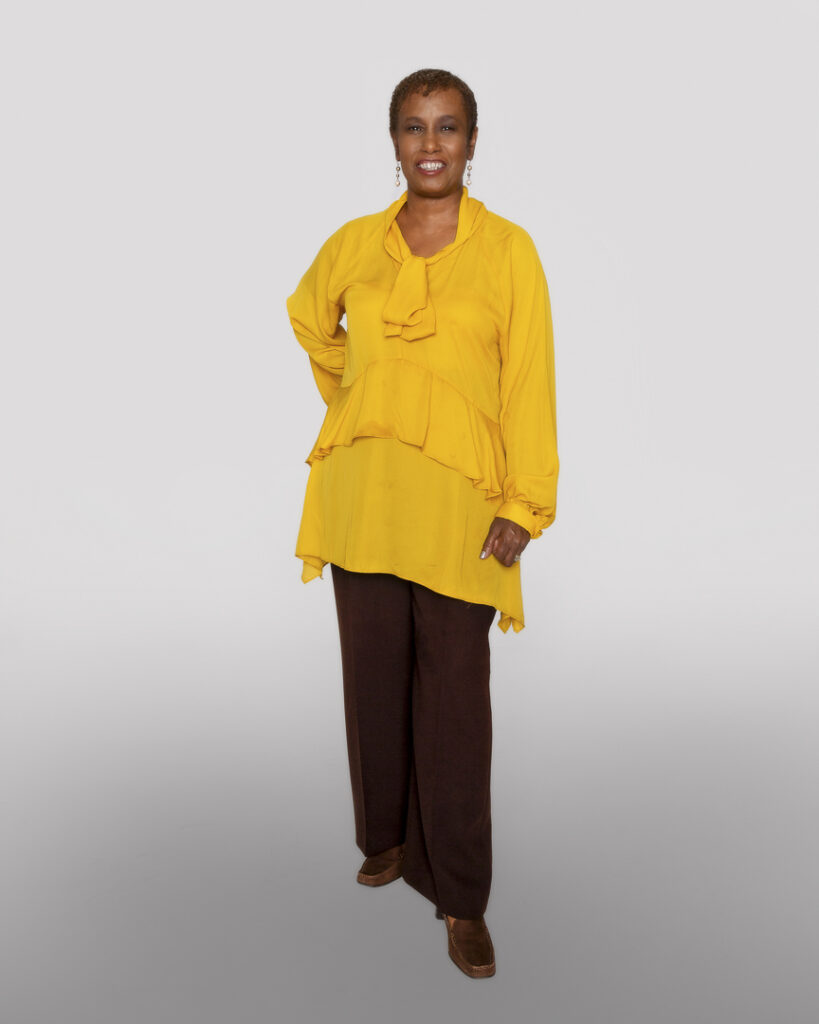 Helen standing in front of a white background wearing black pants and a yellow blouse with fringe